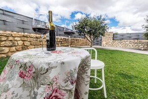 A table with a floral tablecloth, two wine glasses, and a bottle of wine, set outdoors with a stone wall and a tree in the background.