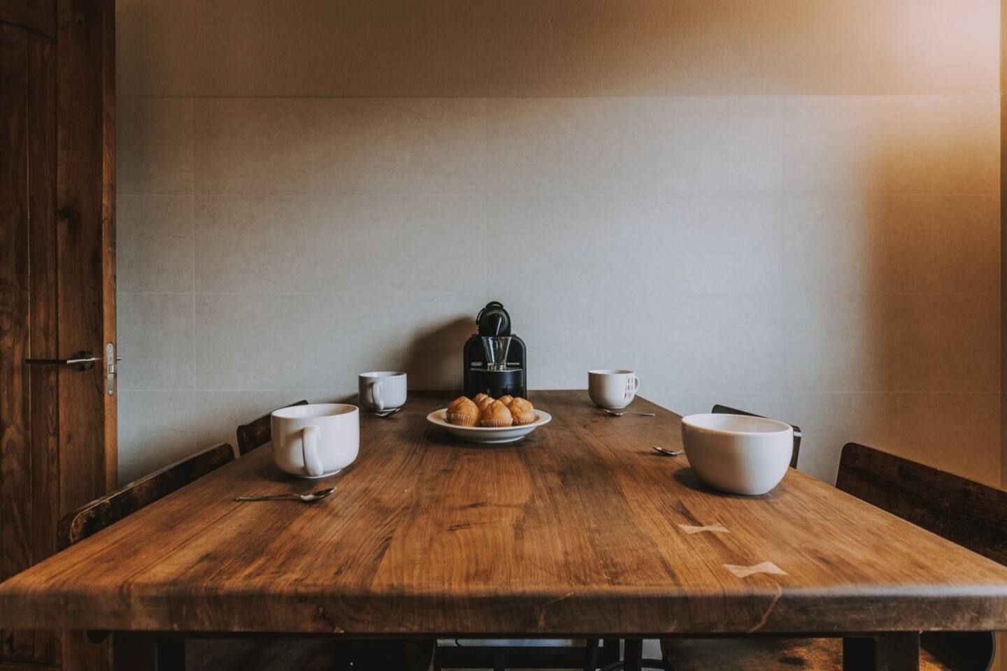 Una mesa de madera con cuatro tazas blancas, un plato de croissants y una cafetera negra.