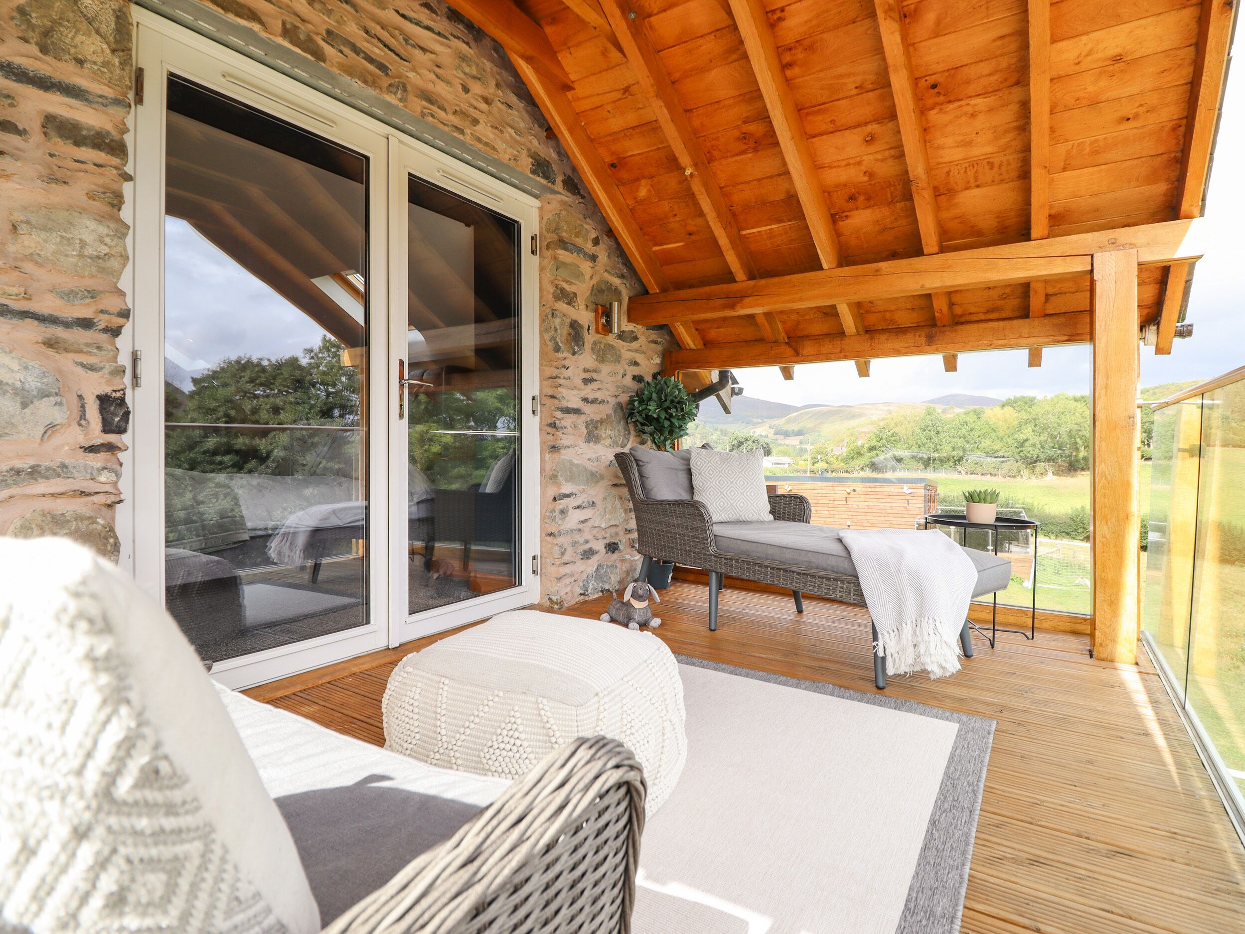 A covered outdoor seating area with wicker furniture, a stone wall, and a view of the countryside.