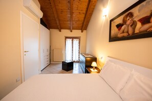 A bedroom with a bed, a nightstand, a lamp, a radiator, a window with curtains, a door, and a painting on the wall.