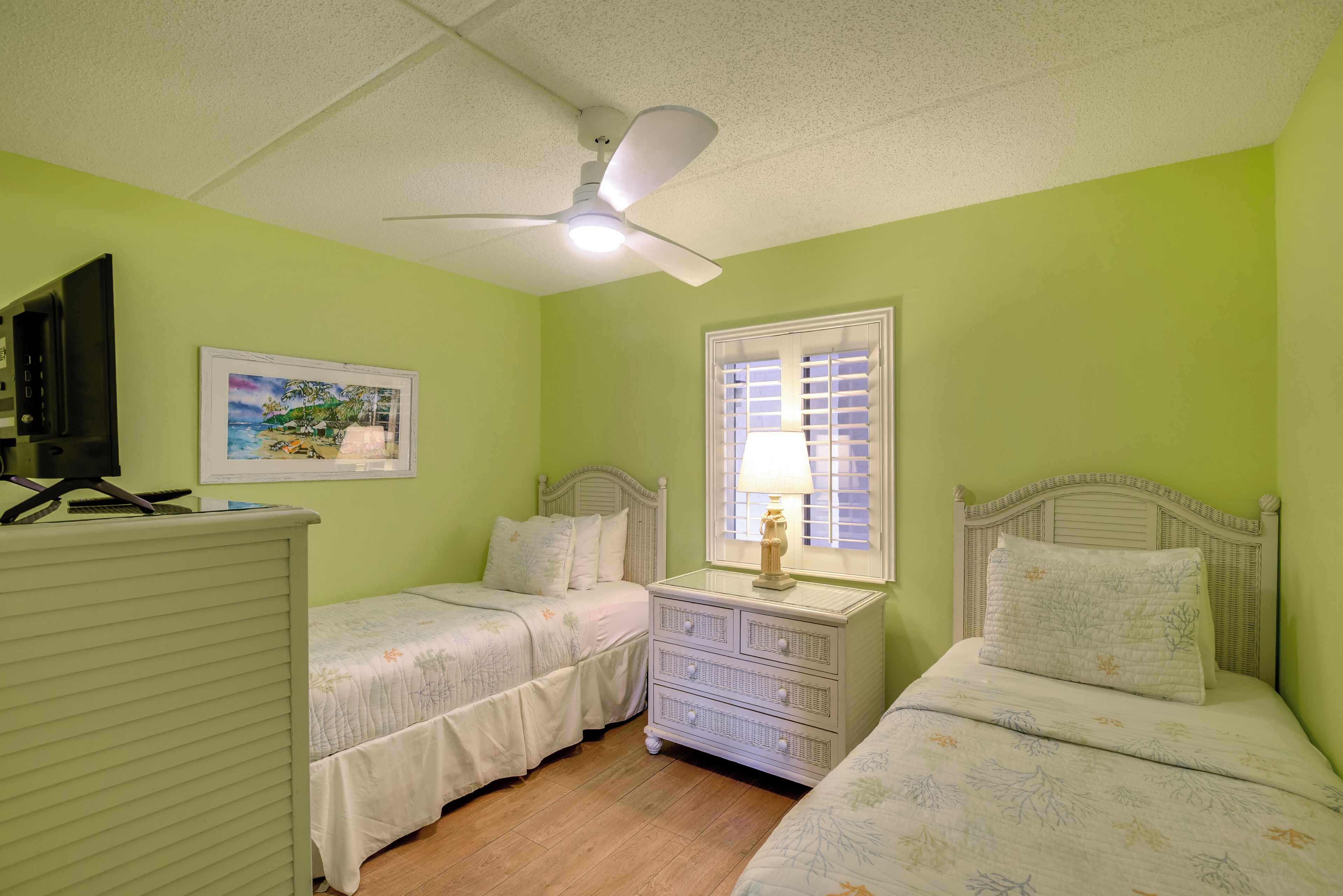 A bedroom with two beds, a nightstand, a desk with a chair, a ceiling fan, and a window with blinds.