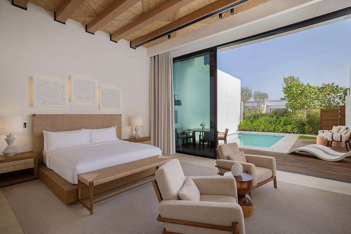 A modern hotel room with a large bed, a seating area with chairs and a bench, a pool view, and artwork on the wall.