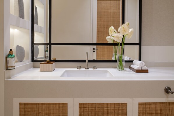 A bathroom with a white sink, a mirror, and a vase with white flowers. - Maysan Doha Lxr Hotels & Resorts (Al Rayyan)