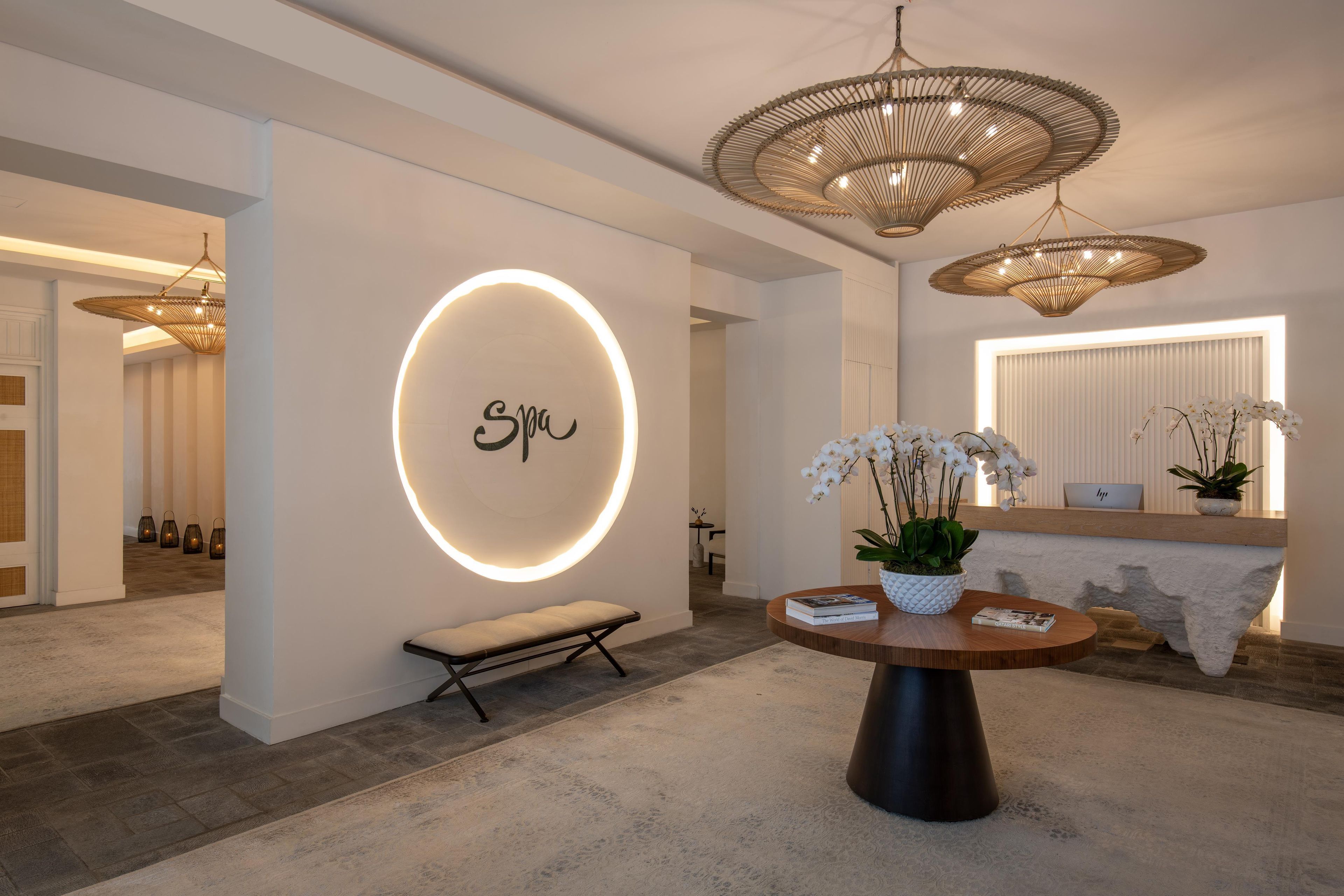 A modern interior with a circular illuminated wall sign, a bench, a round table with books and a potted plant, and a large woven light fixture.