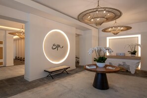 Un intérieur moderne avec un panneau mural circulaire rétroéclairé, un banc, une table ronde avec des livres et une plante en pot, ainsi qu’un grand luminaire tissé.