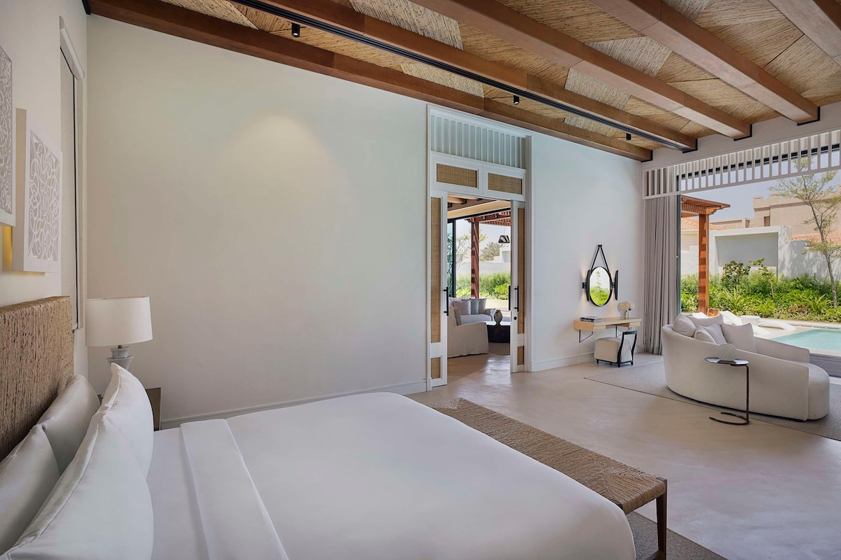 A modern hotel room with a large bed, a sofa, a small table, and a view of the outdoors.
