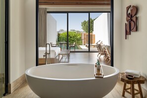 A modern bathroom with a large freestanding bathtub, a wooden stool with a towel, and a view of an outdoor patio with a pool and furniture. - Maysan Doha Lxr Hotels & Resorts (Al Rayyan)