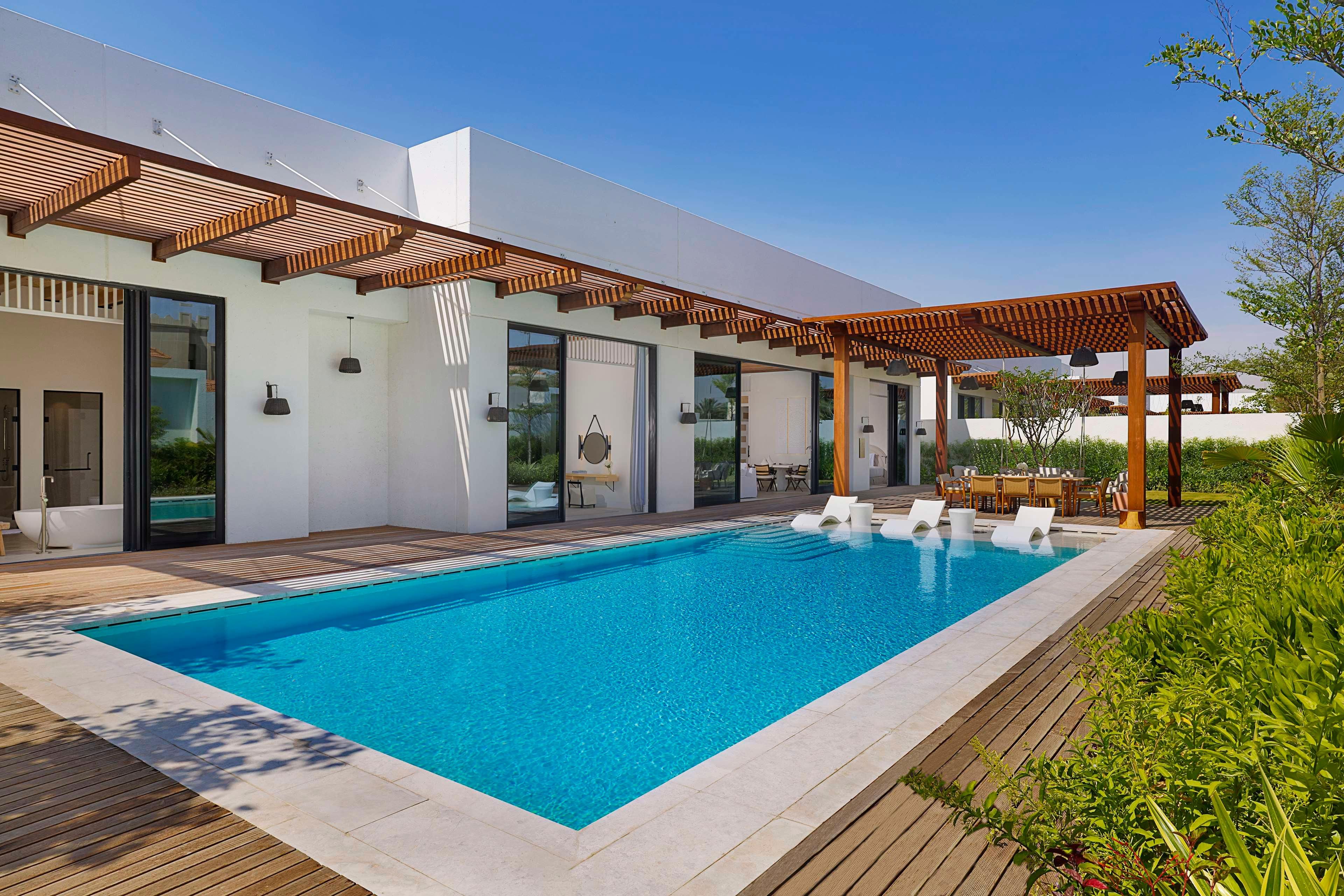 Een modern buitenzwembad met een houten terras, een pergola en een eethoek met tuinmeubilair.