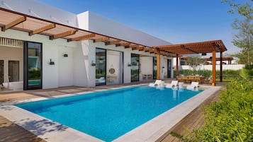 Een modern buitenzwembad met een houten terras, een pergola en een eethoek met tuinmeubilair.