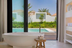 Une salle de bain moderne avec une baignoire sur pieds, un petit tabouret en bois et une vue sur une piscine et la verdure environnante.