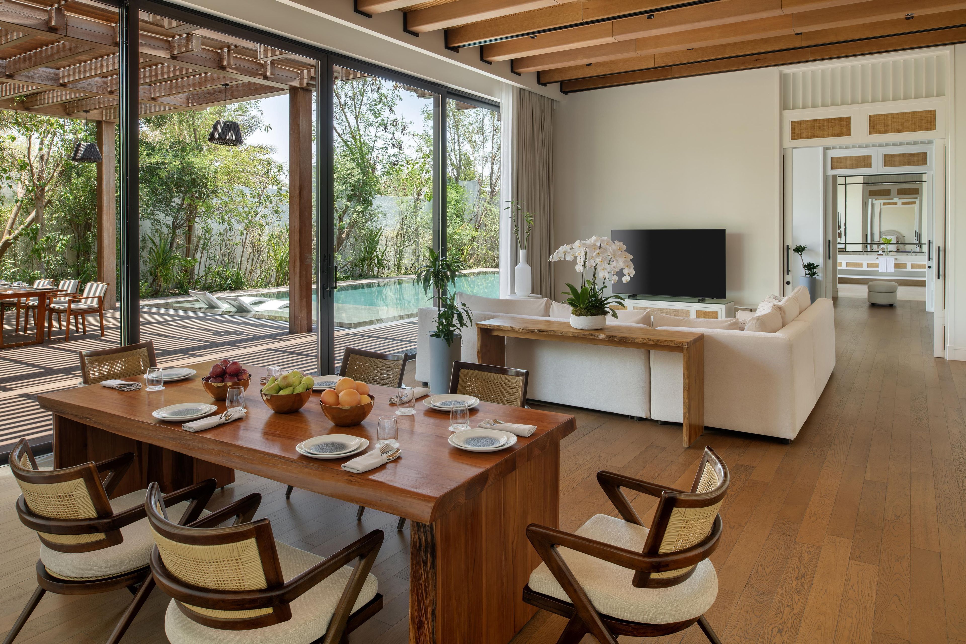 A modern living room with a wooden dining table set for four, a sofa, a TV, and a view of a pool and greenery.