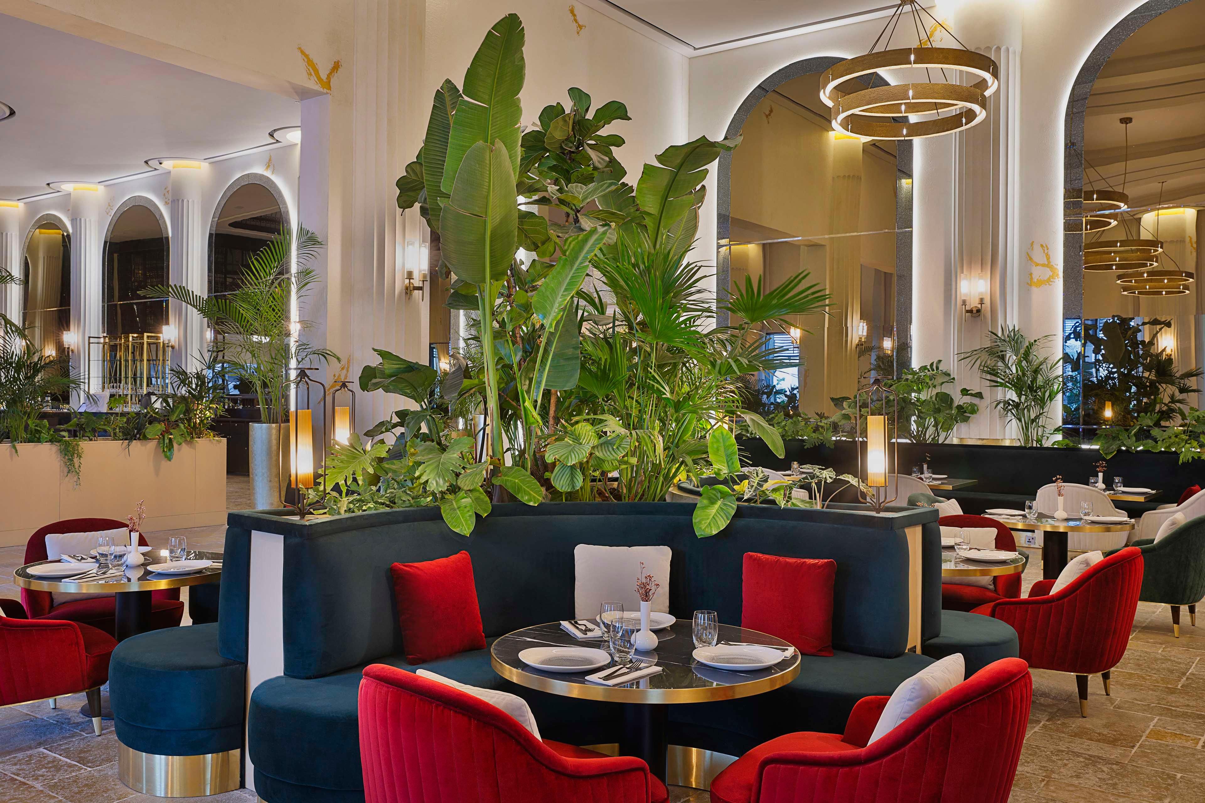 Un ristorante con tavoli rotondi, sedute imbottite rosse e blu, e grandi piante verdi.