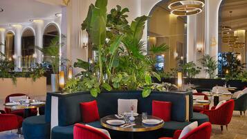 Un ristorante con tavoli rotondi, sedute imbottite rosse e blu, e grandi piante verdi.