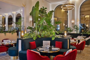 A restaurant with round tables, red and blue upholstered seating, and large green plants. - Maysan Doha Lxr Hotels & Resorts (Al Rayyan)