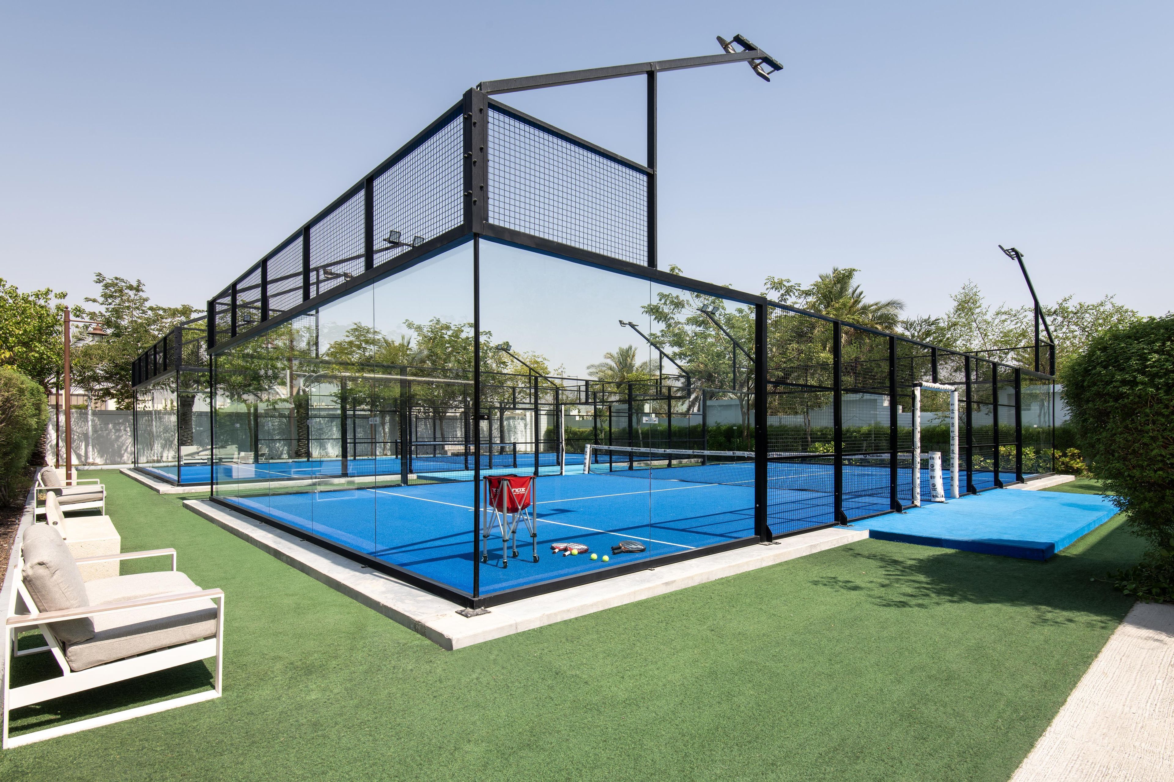 A tennis court with a net, surrounded by a metal fence, and a seating area with chairs.