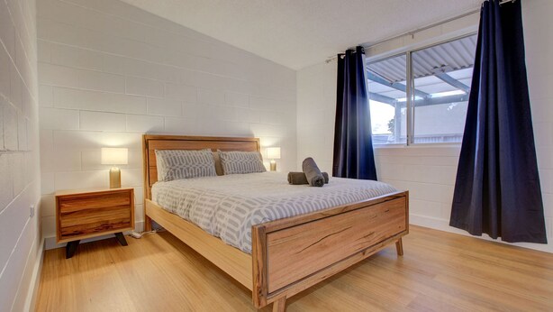 A bedroom with a wooden bed, two bedside tables with lamps, a window with dark curtains, and a white brick wall.