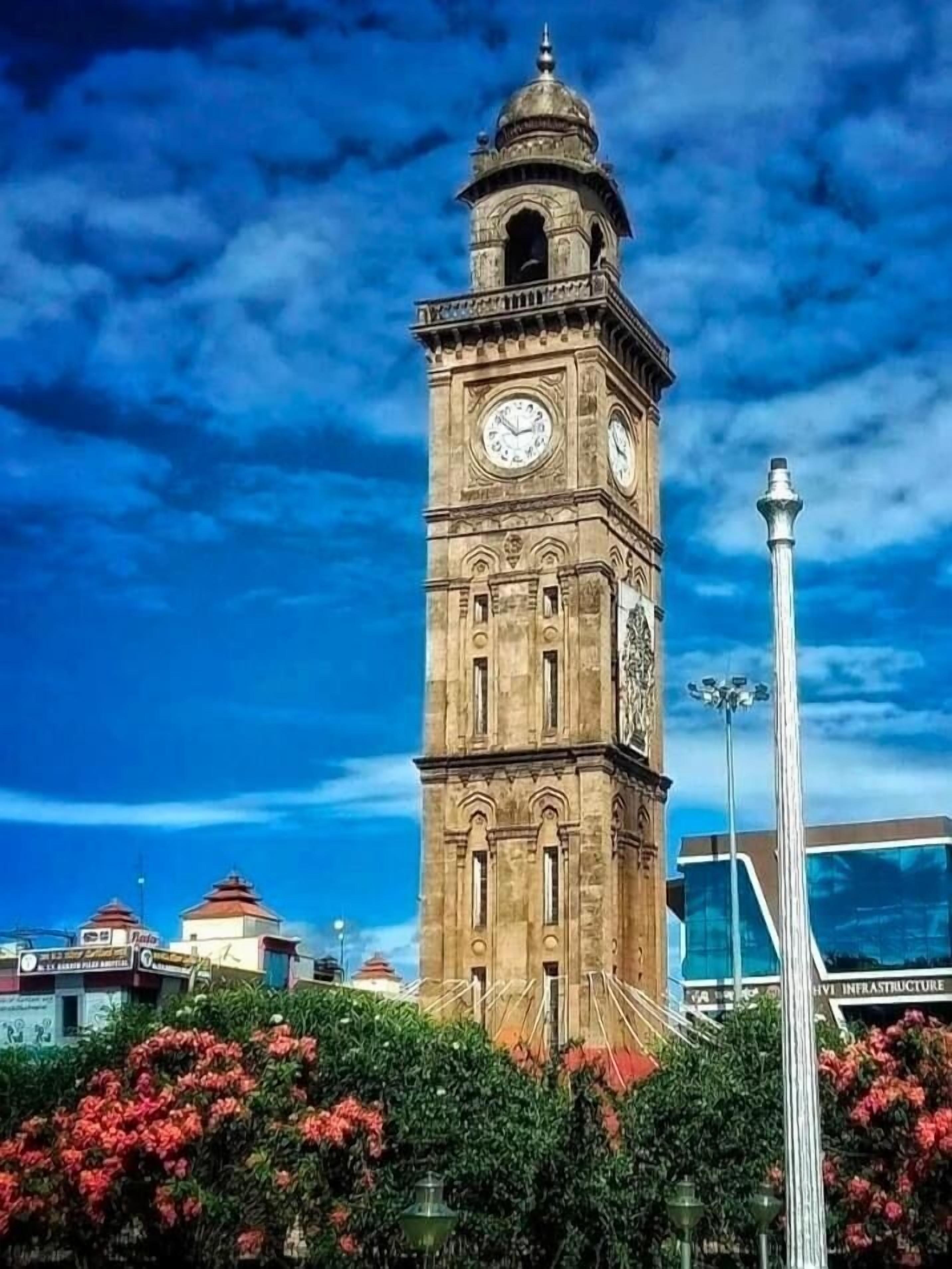 A historic clock tower with a dome and two clocks, surrounded by modern buildings and flowering bushes.