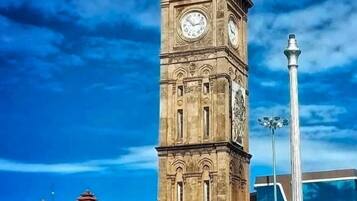 A historic clock tower with a dome and two clocks, surrounded by modern buildings and flowering bushes.