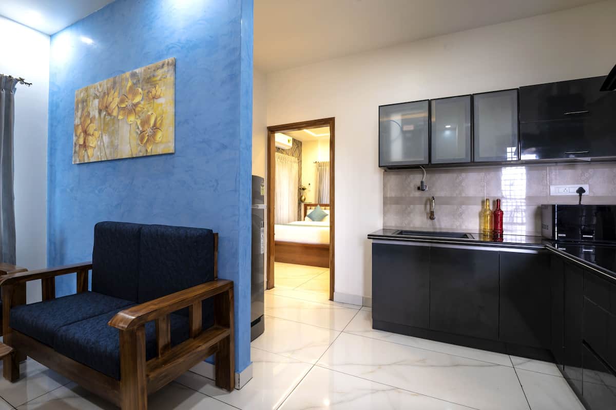 A compact living space with a blue accent wall, a wooden sofa, a small dining table, a kitchen with black cabinets and a microwave, and a painting on the wall.