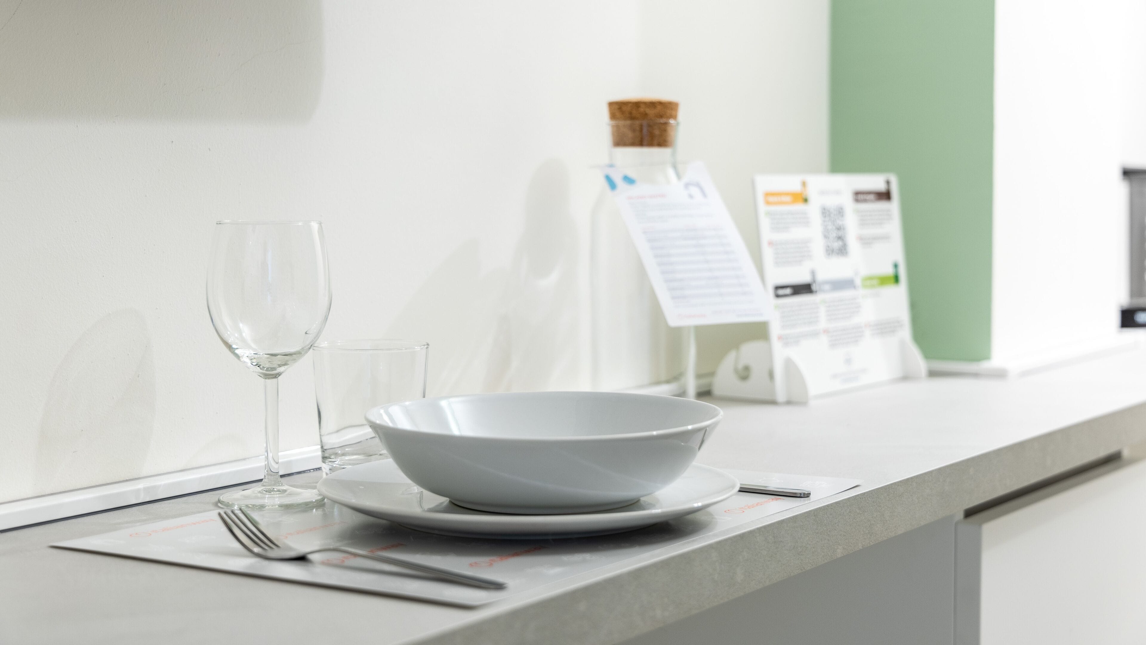 Un comedor moderno y luminoso con una mesa blanca dispuesta para dos personas, incluyendo un bol, platos y cubiertos. Se ve también una copa de vino y una botella de agua.