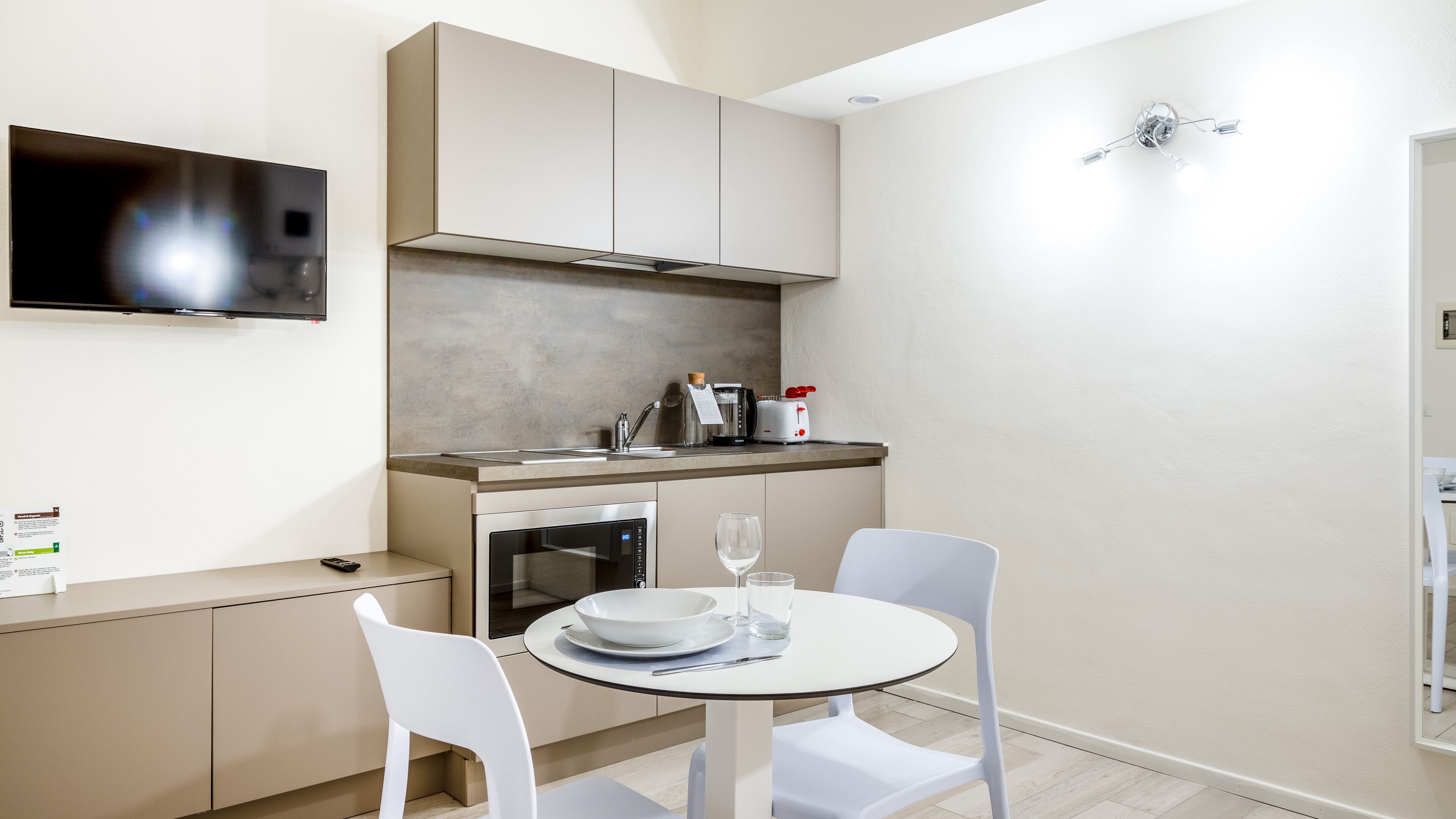 A compact kitchen with modern appliances, a dining area with white chairs, and a flat-screen TV mounted on the wall.