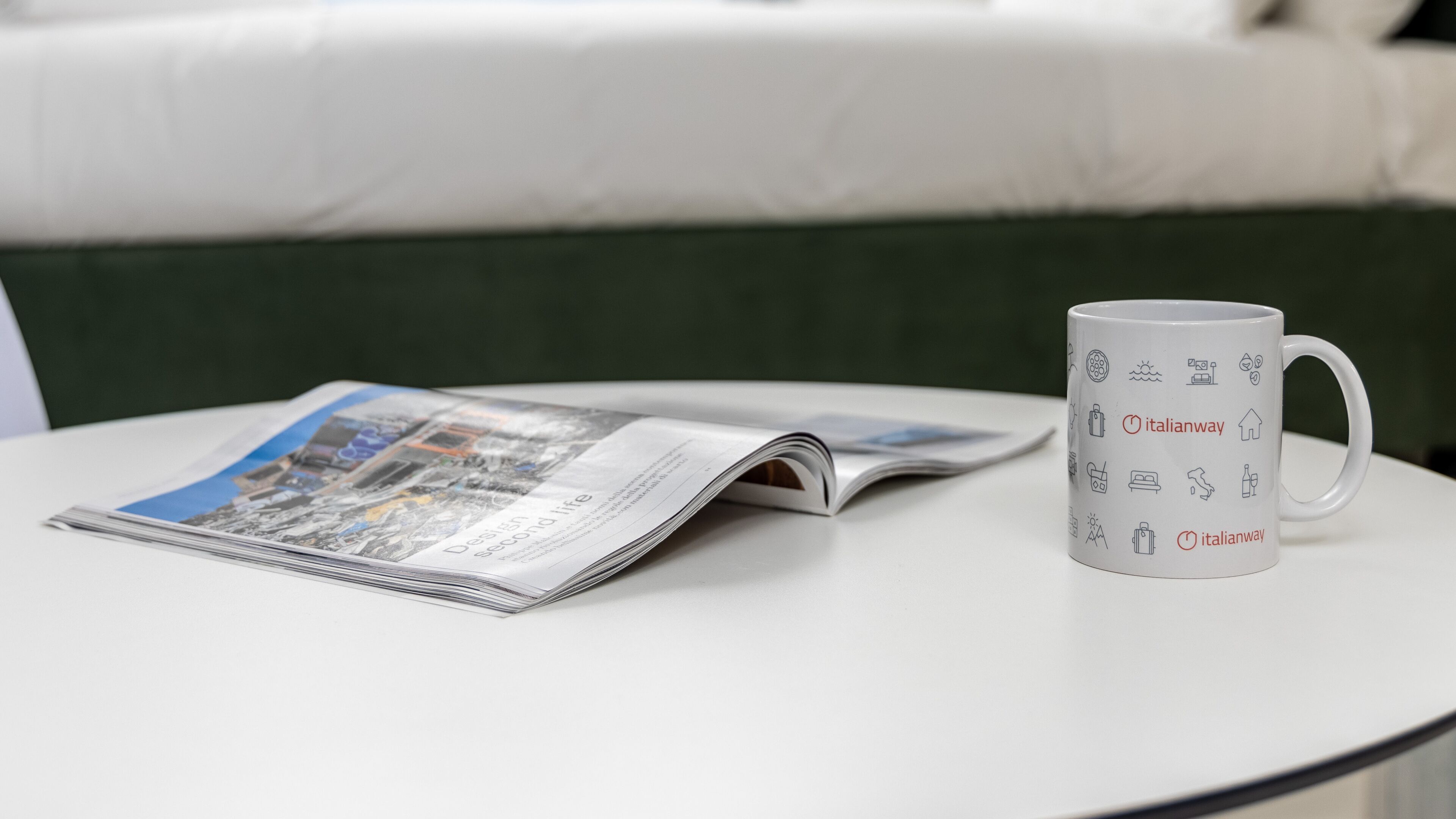 Eine weiße Tasse mit Italianway-Branding auf einem weißen Tisch, neben einer geöffneten Zeitschrift mit Stadtansicht.