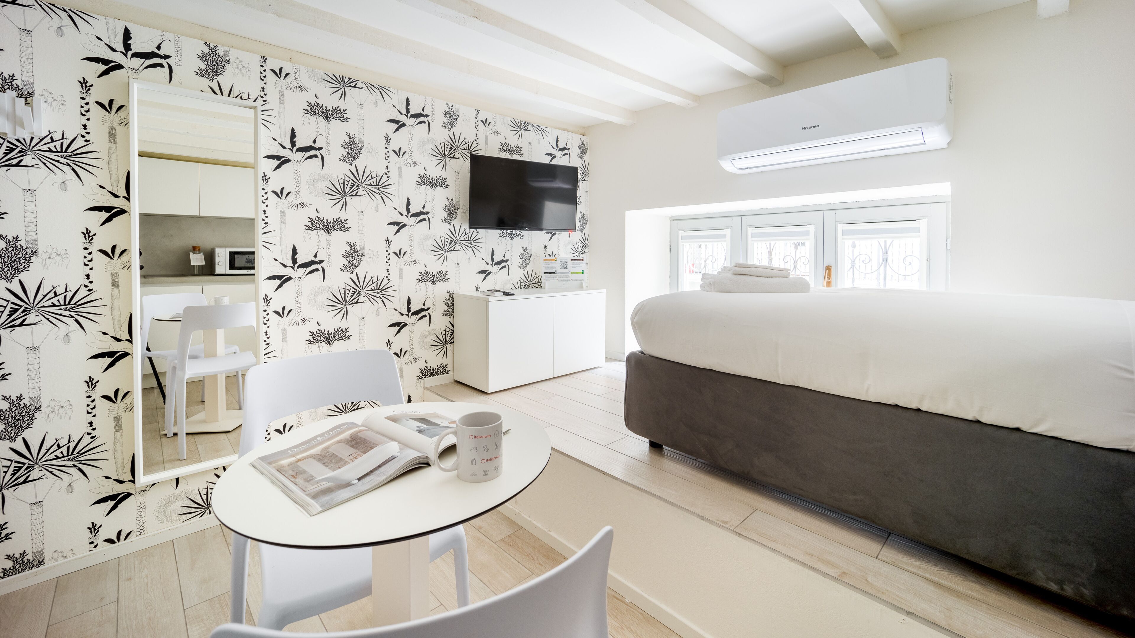 A modern hotel room with a bed, a TV, a dining area with a table and chairs, and a bathroom visible through an open door.