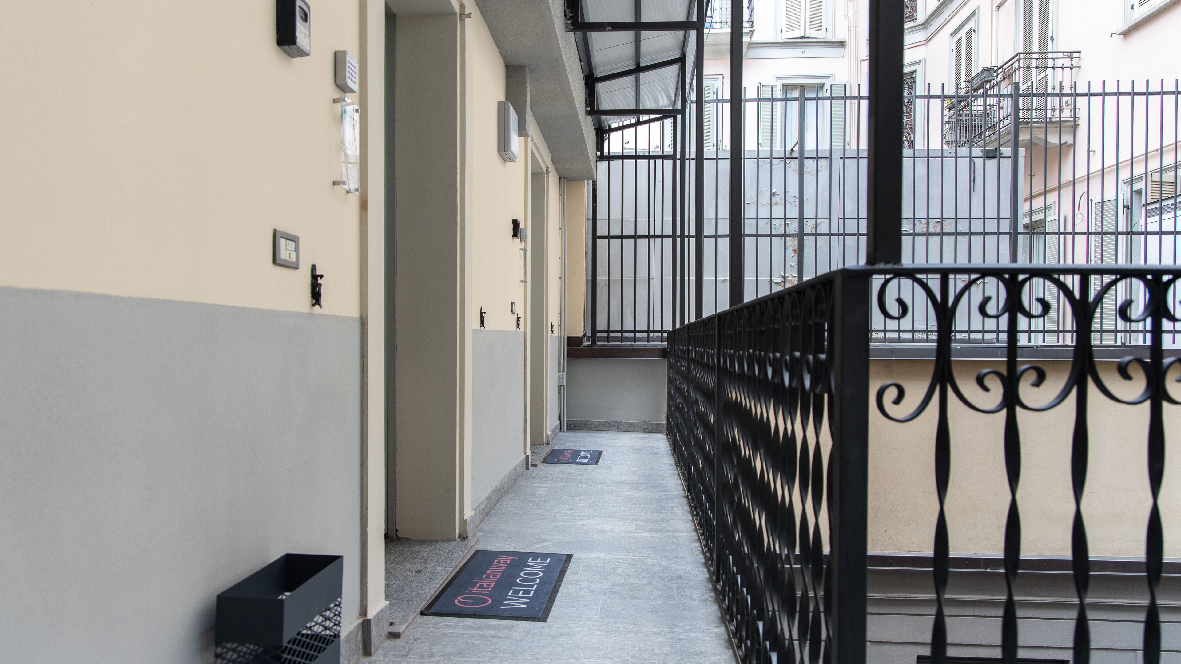 Un couloir étroit avec une rambarde en métal, un paillasson et une porte donnant sur un balcon.