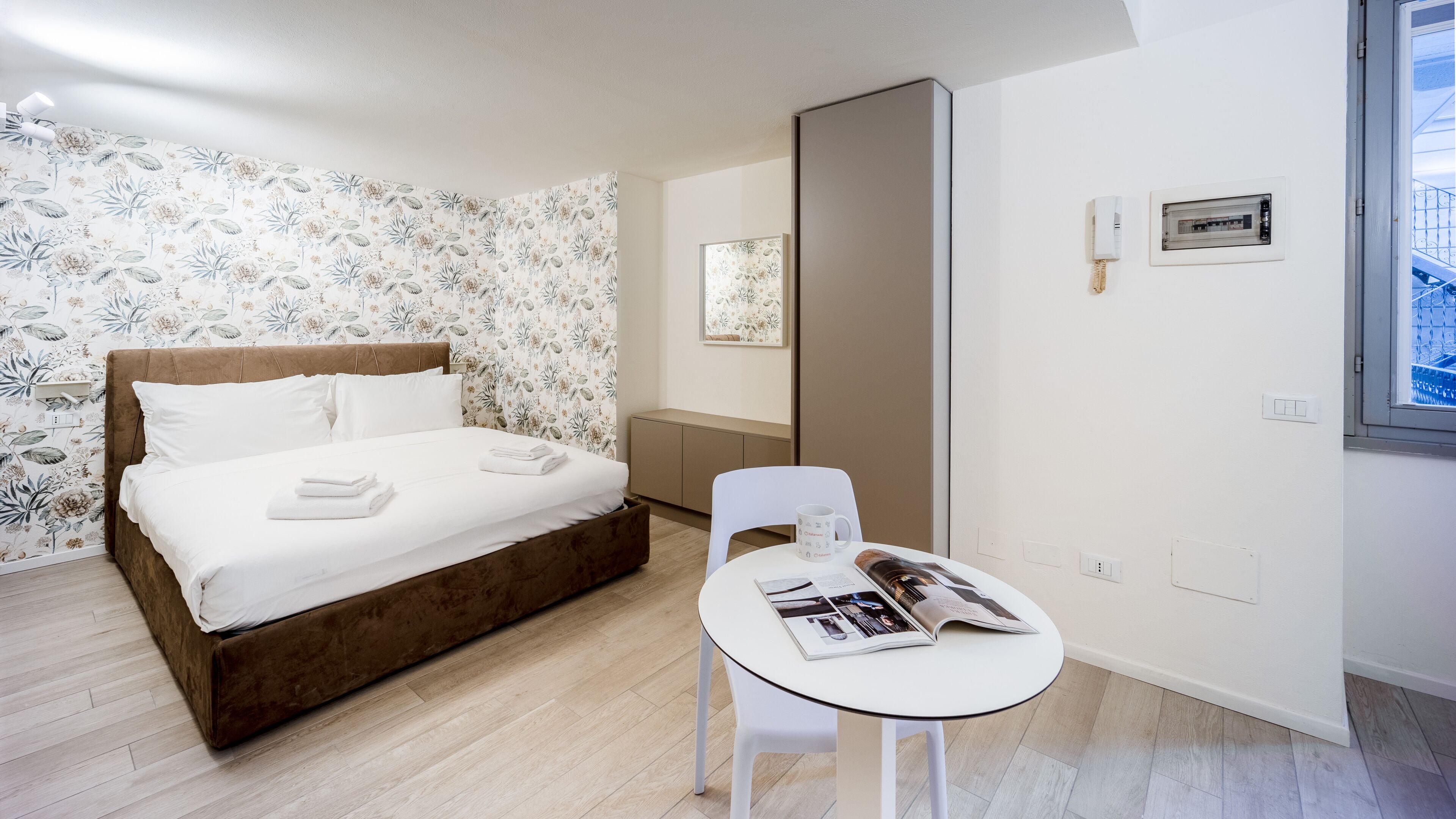 Un dormitorio ordenado con una cama, una mesita de noche con una revista, una silla y un ventanal con vistas.