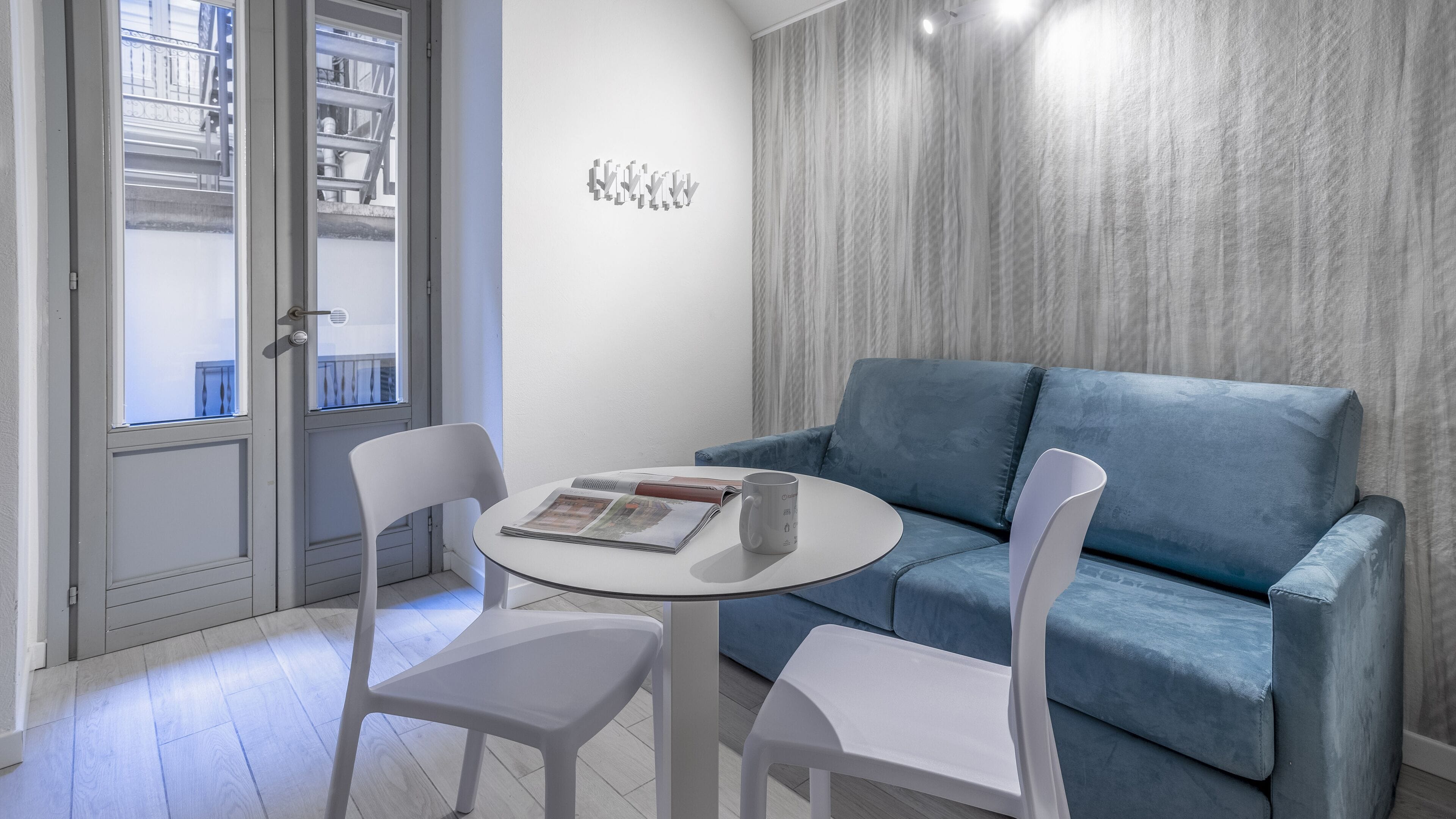 Un salon moderne avec un canapé bleu, deux fauteuils blancs, une table ronde sur laquelle reposent un magazine et une tasse, et une vue à travers une porte vitrée.