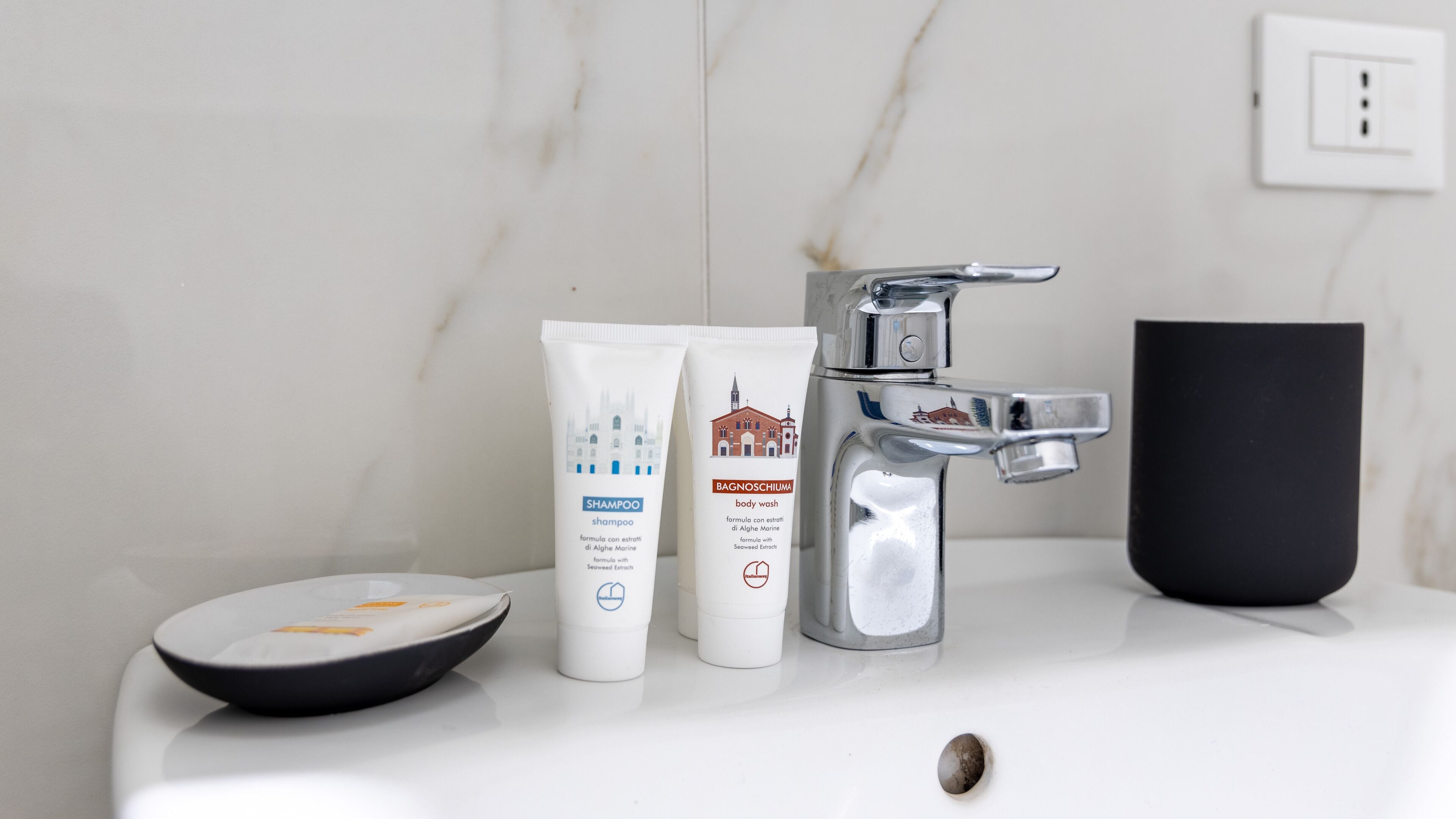 A bathroom sink with a marble wall, a black mug, and two toiletry bottles.