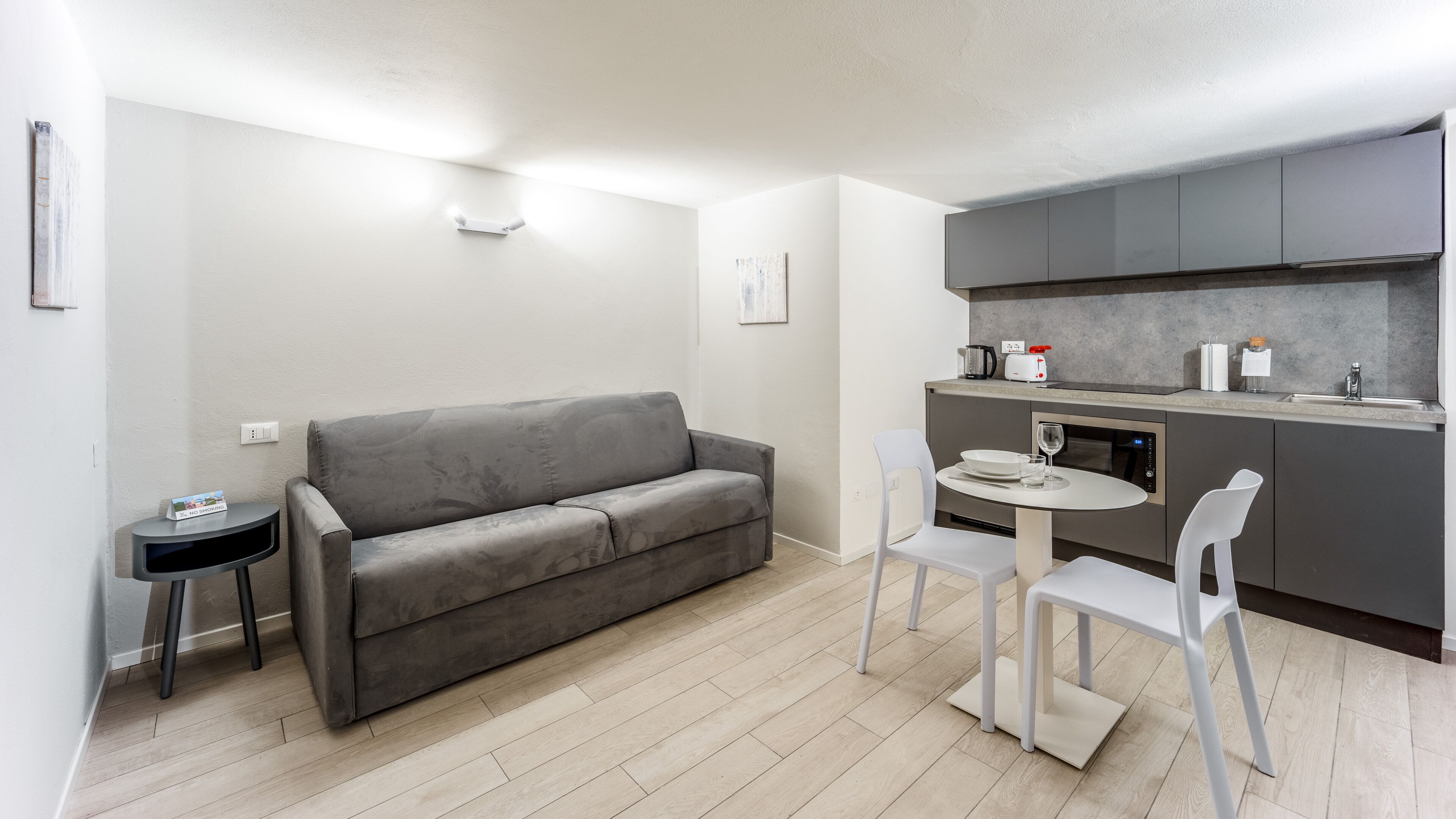 A compact living space with a grey sofa, a small round table, and two white chairs. A kitchenette with grey cabinets and a countertop is visible.