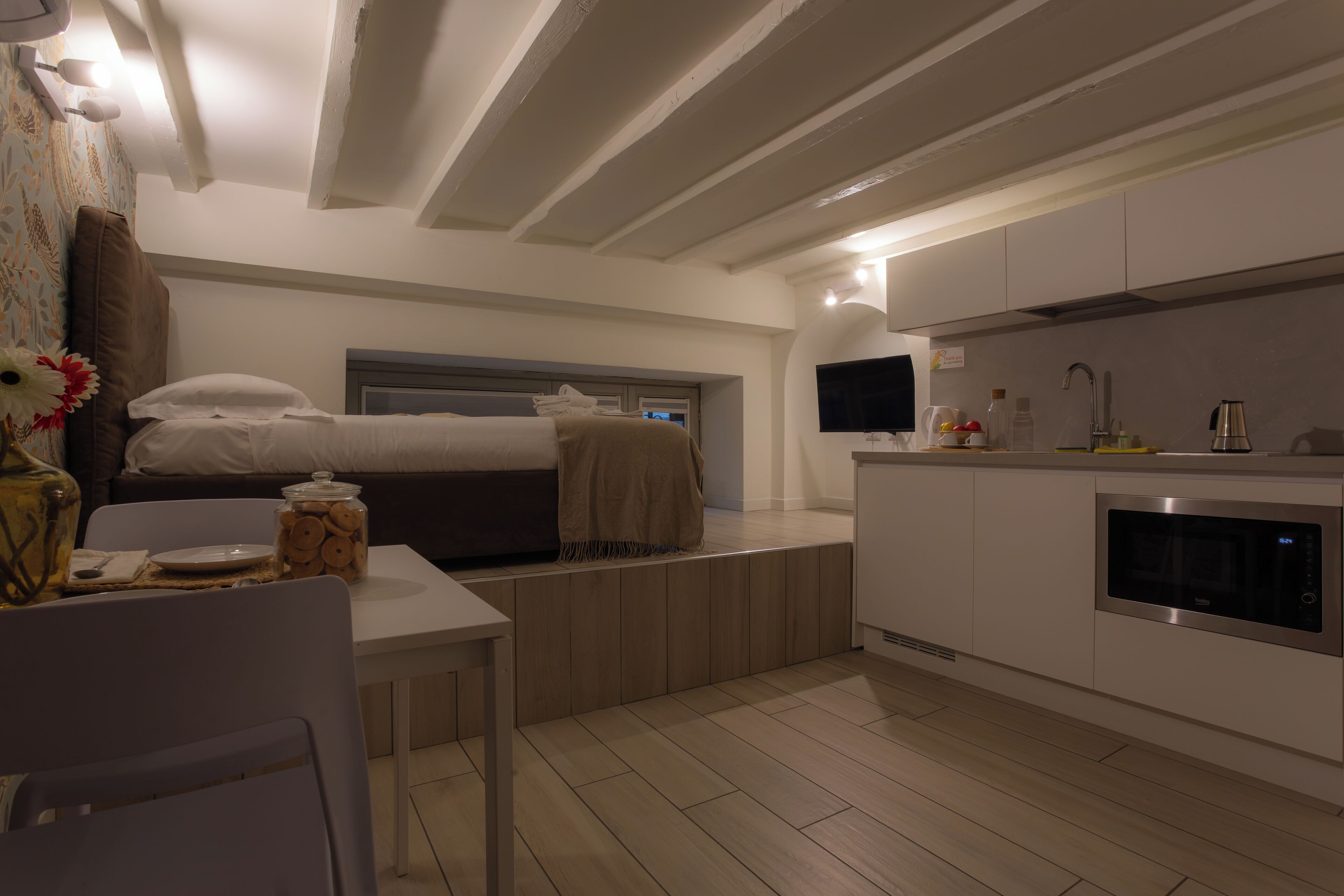 A compact bedroom with a bed, bedside table, and a small table with a vase and jar. Adjacent is a kitchenette with a microwave, sink, and countertop.