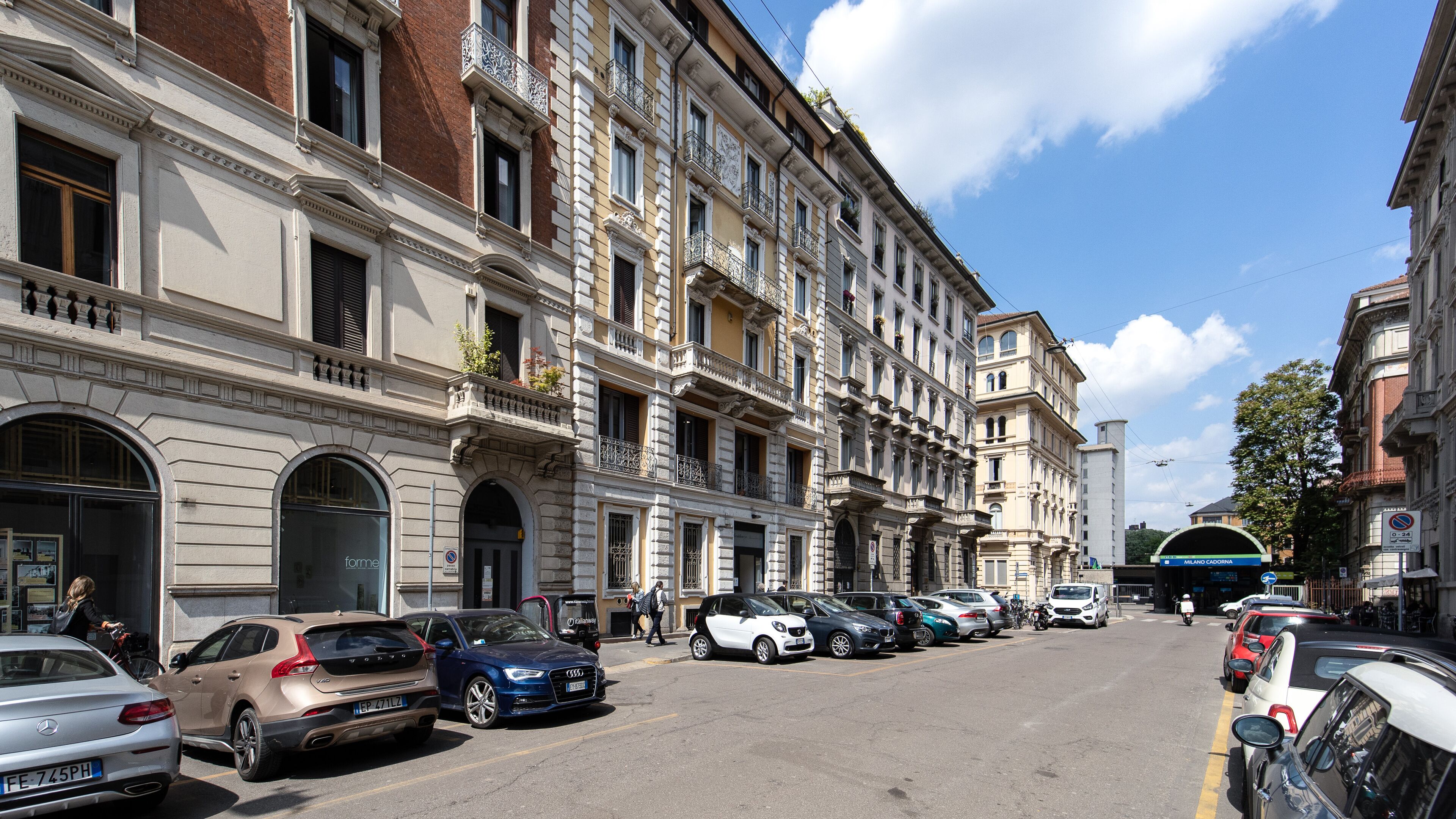 Una calle flanqueada por edificios de varios pisos, coches aparcados y un cielo azul despejado.