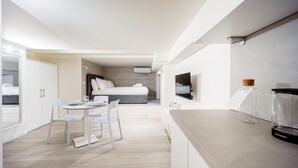 Quarto de hotel moderno com cama, pequena área de refeições com mesa e cadeiras, televisão na parede e minibar.