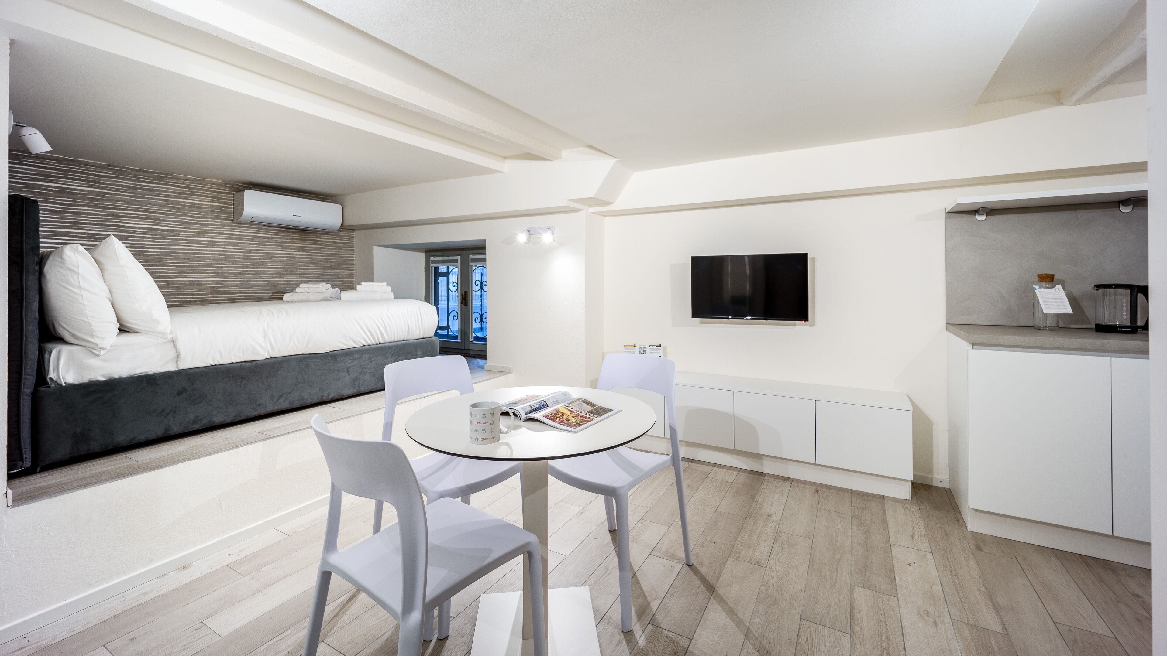 A modern hotel room with a bed, a dining area with a table and chairs, a TV mounted on the wall, and a kitchen area with a coffee maker.