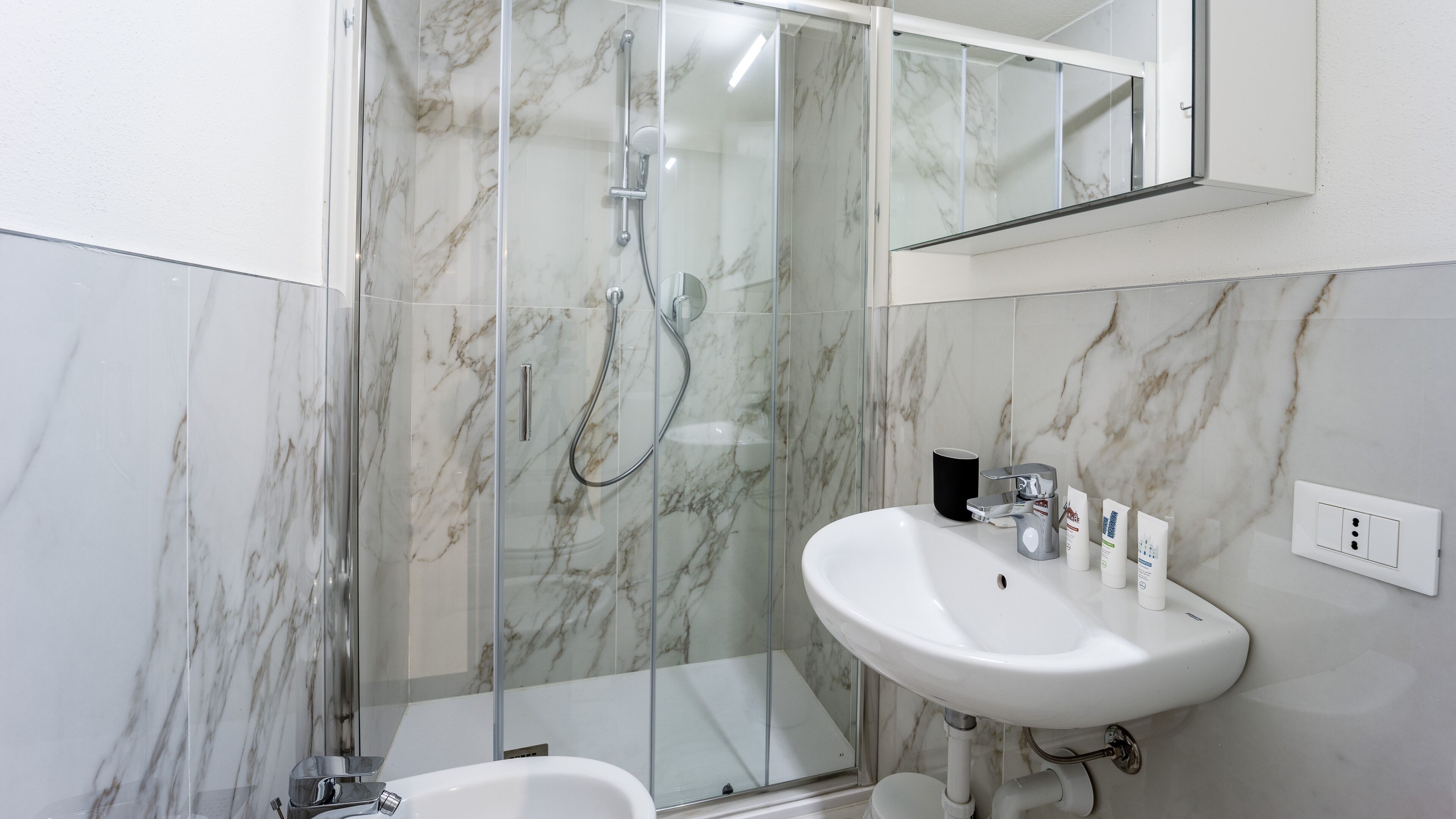 Un baño con ducha envuelta en vidrio, lavamanos blanco y paredes de mármol.