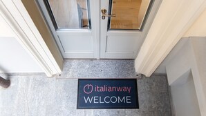 Uma porta com painel de vidro e puxador de latão, tapete de entrada com a inscrição 'Italianway WELCOME' e piso de ladrilho de pedra.