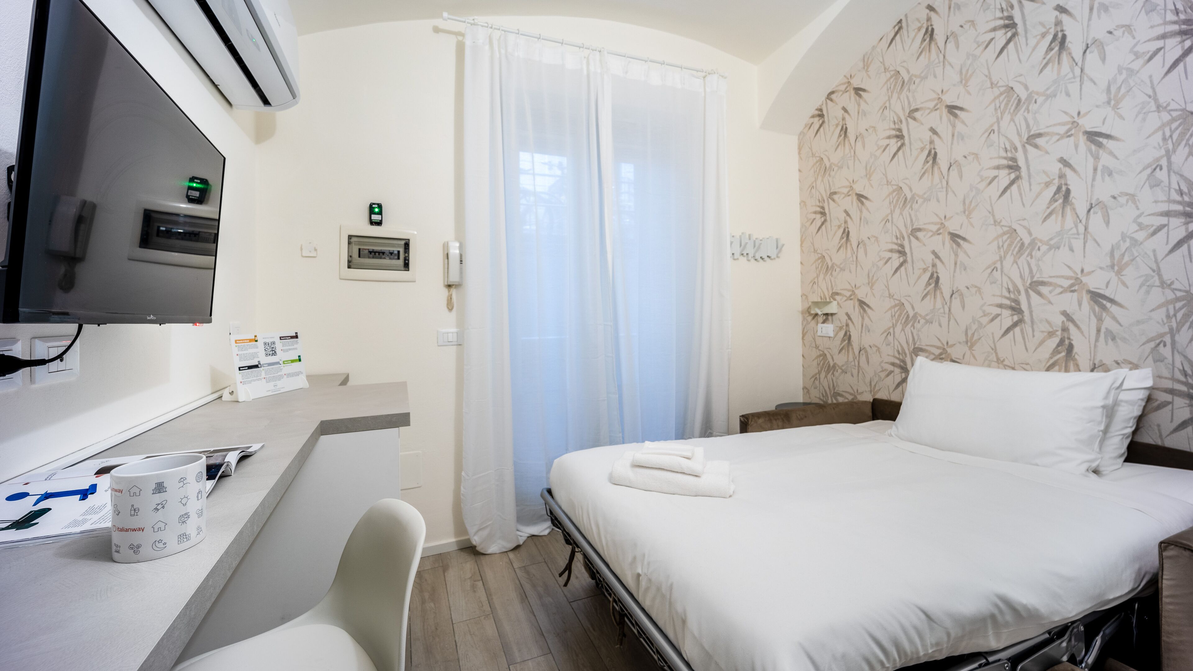 Una habitació d'hotel amb llit, escriptori amb ordinador, cadira i finestra amb cortines.