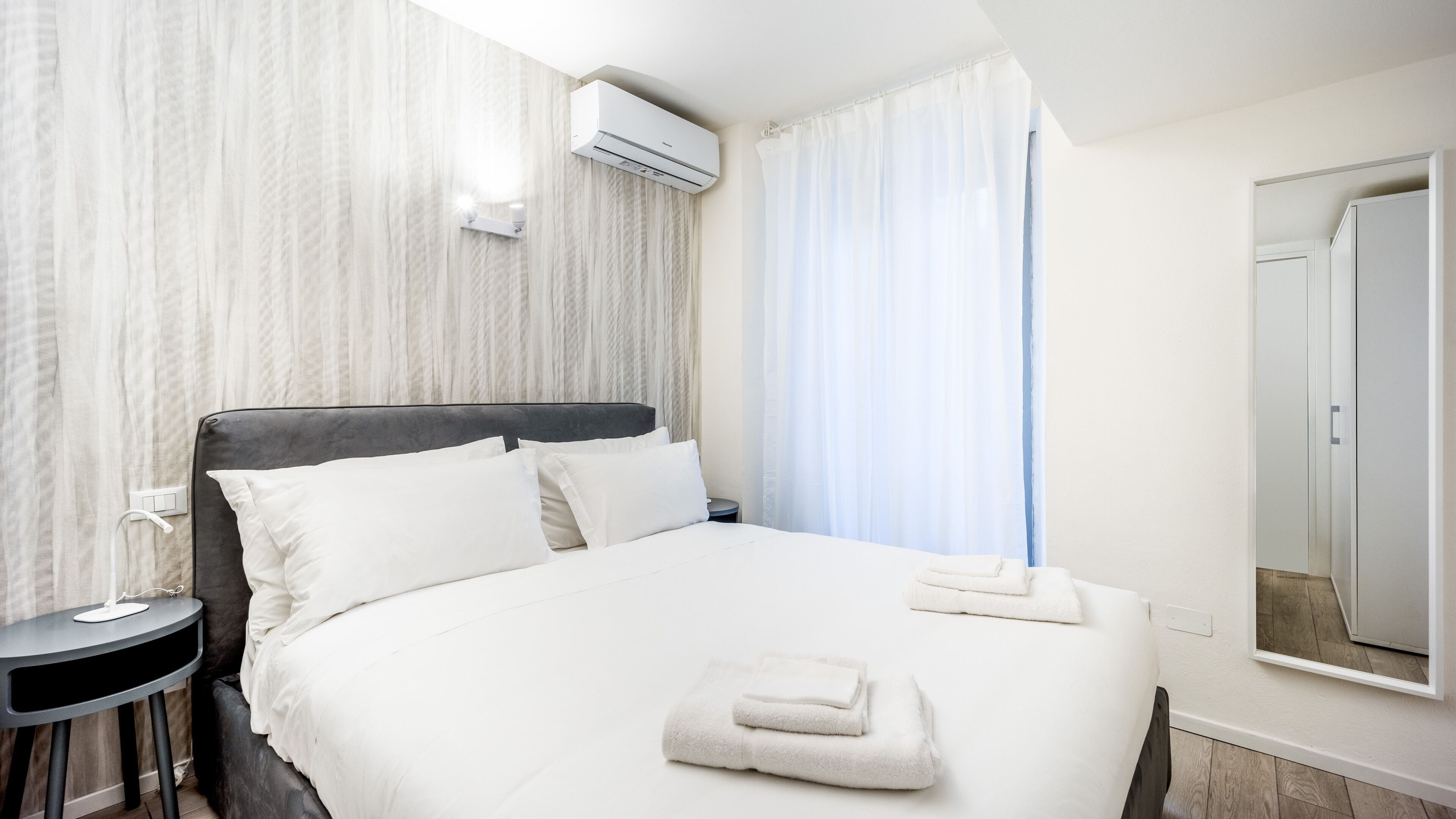 A hotel room with a bed, a bedside table, an air conditioning unit, a mirror, and a window with curtains.