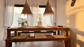 Un espace repas avec une table et un banc en bois, des rideaux blancs et des luminaires suspendus en fibres tressées.
