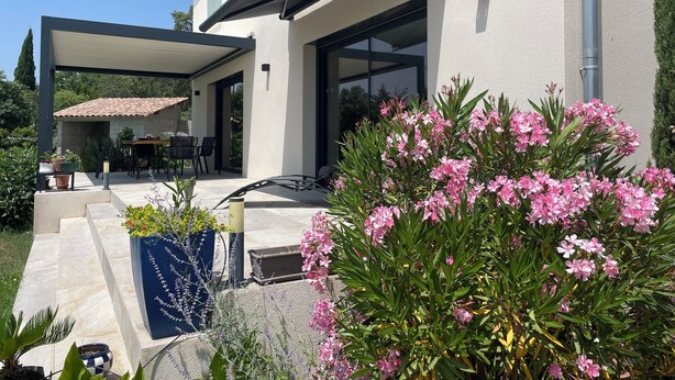 Una casa moderna con porche cubierto, mobiliario exterior y un jardín con flores rosadas.