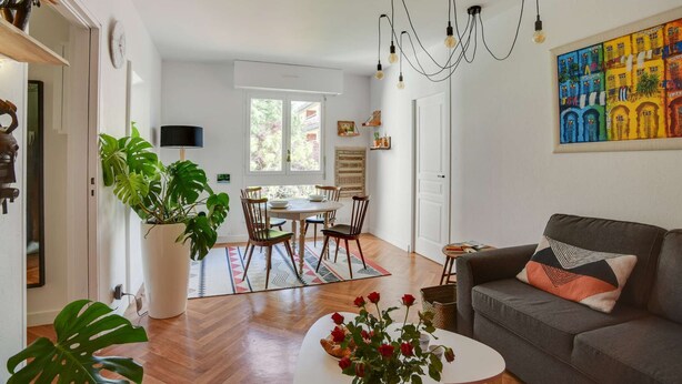 Un salon comprenant un canapé, une table basse et un coin repas avec des chaises et une table. On y trouve également des plantes en pot, un tableau accroché au mur et un lustre.