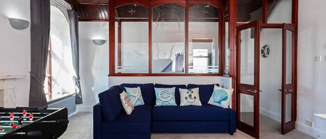 A room with a foosball table, a blue sofa with decorative pillows, and a large window with a view of the outside.