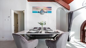 A dining area with a white table set for two, two grey upholstered chairs, a vase with flowers, and a sunset painting on the wall. - Cozy Cove Chapel (Portland)