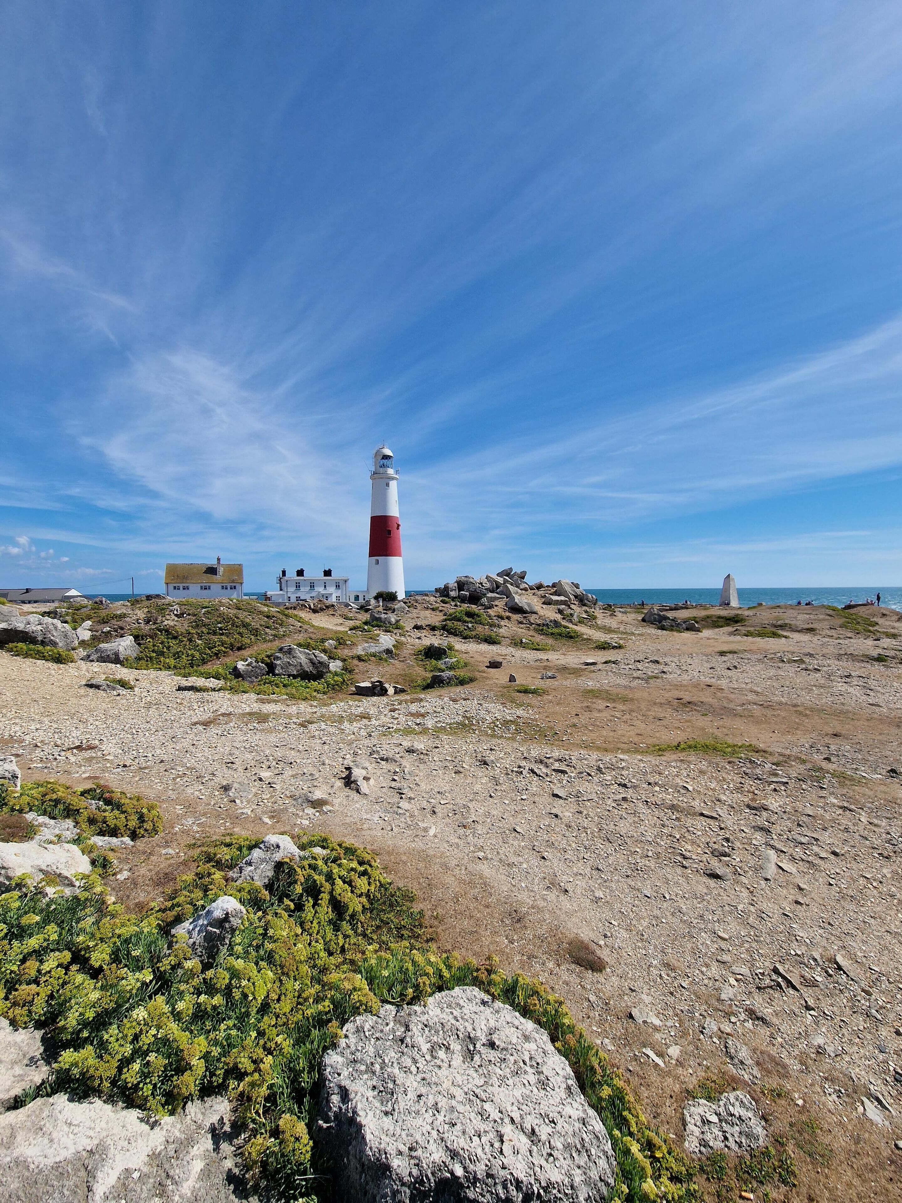 A coastal landscape with a prominent lighthouse, rocky terrain, sparse vegetation, and a clear blue sky.