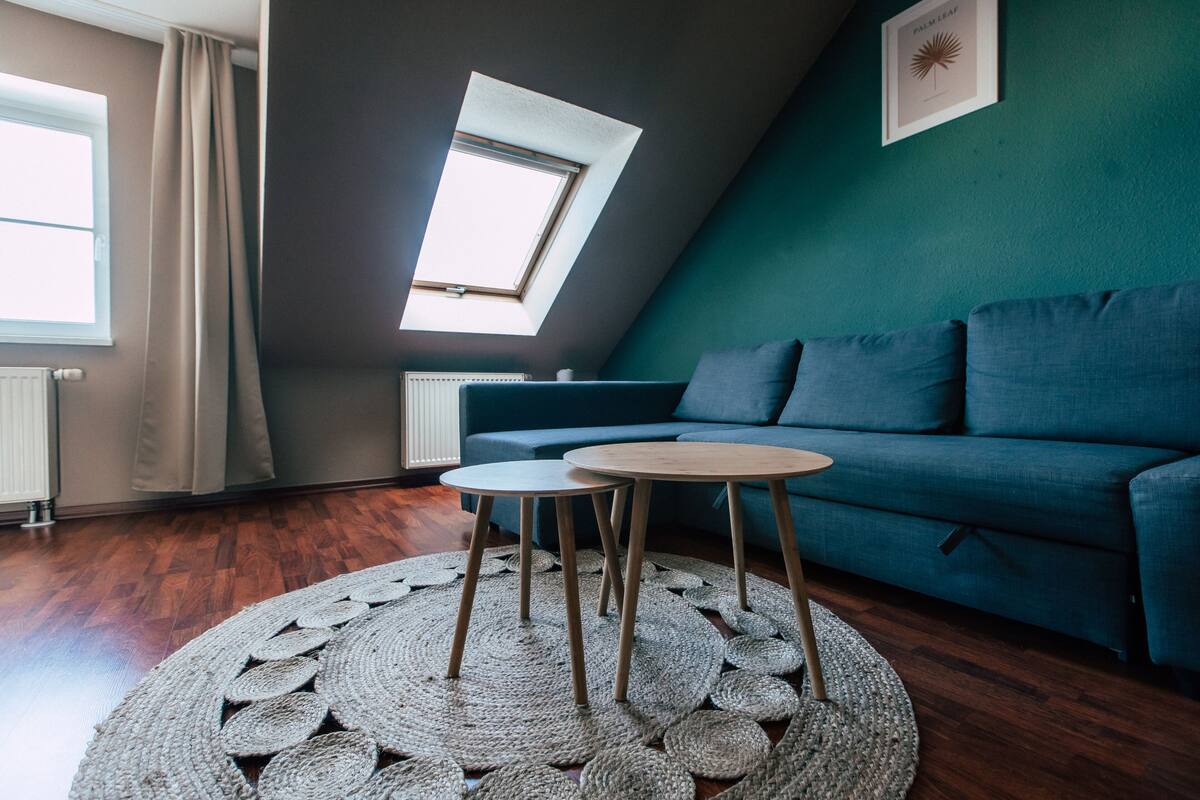 A modern loft bedroom with a sloped ceiling, a skylight, a blue sofa, two round wooden tables, and a woven rug.