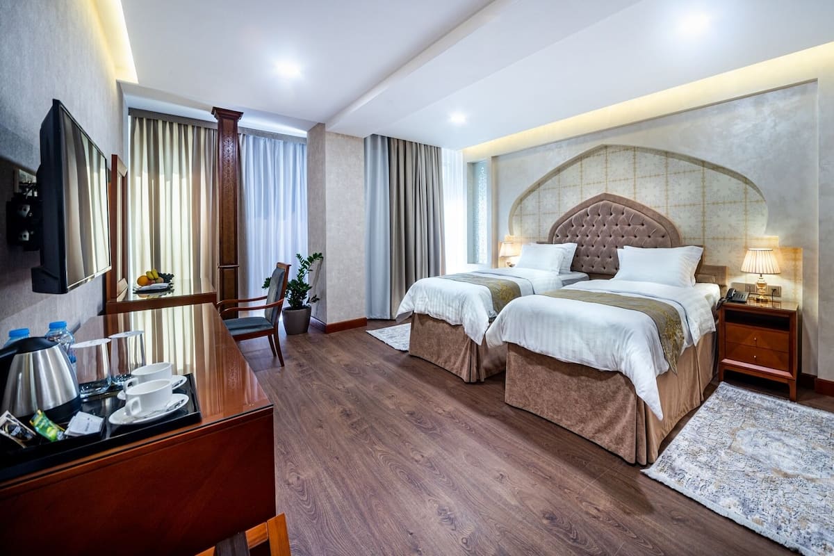 A hotel room with two beds, a desk with a chair, a TV, and a large window with curtains.