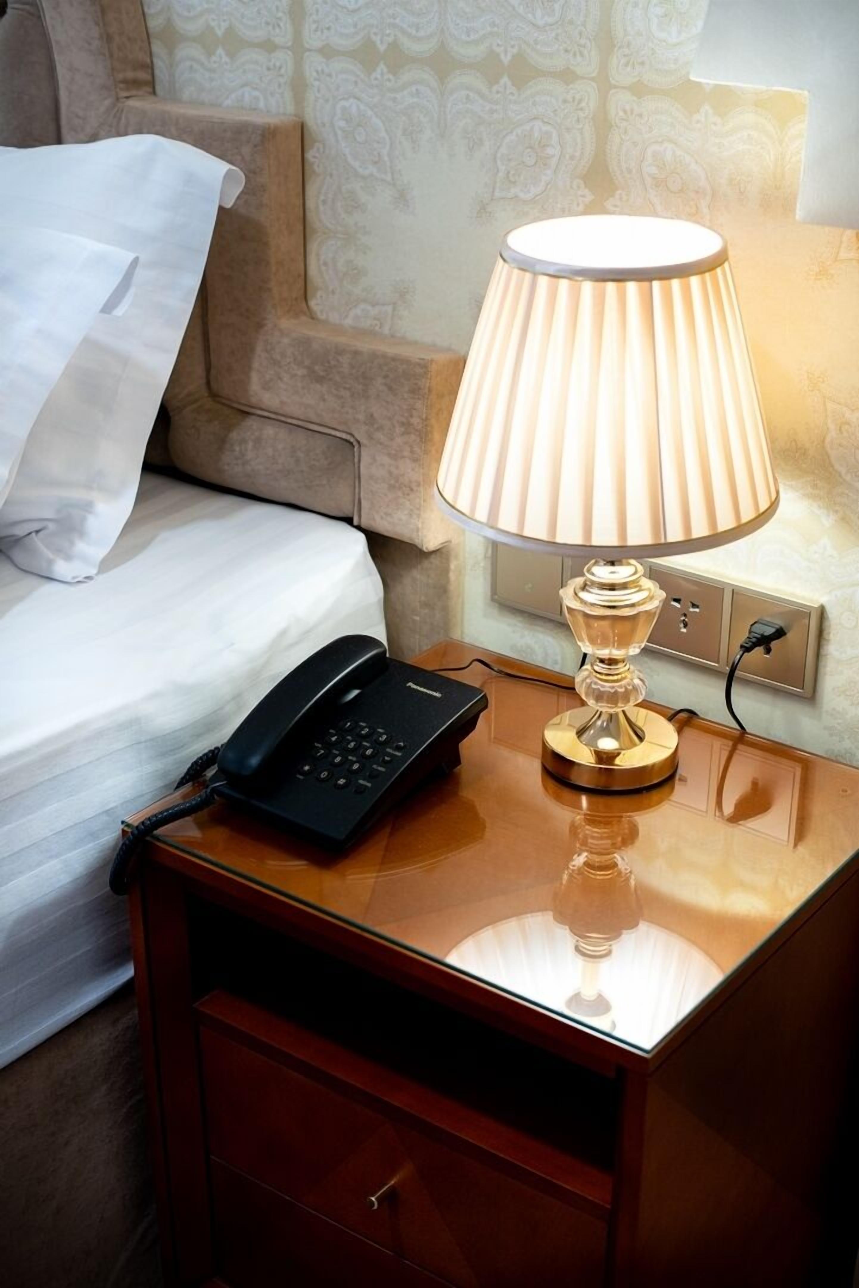 A neatly made bed with a white pillow and a black telephone on a wooden nightstand. A table lamp with a white shade is placed on the nightstand.