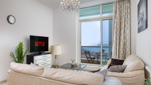 A modern living room with a sofa, a TV displaying 'NETFLIX', a chandelier, a table with a vase, and a balcony view.
